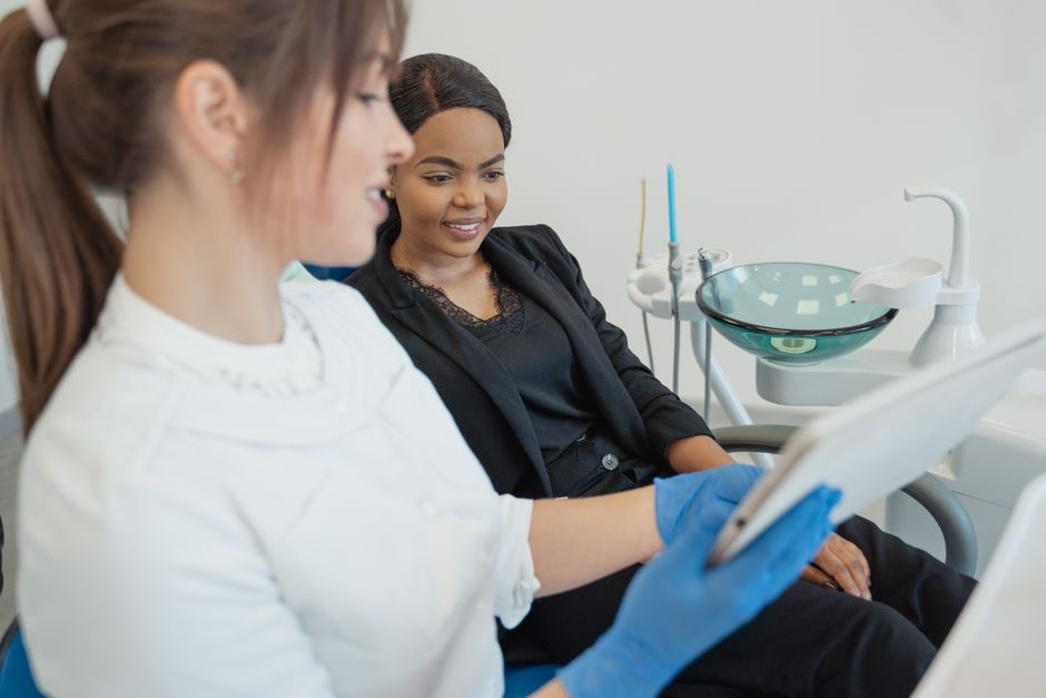 A woman with dark hair and medium skin tone, dressed in a black blazer and lace top, is seated in a dental clinic chair, smiling and engaging with a dental professional. The dental professional, weari