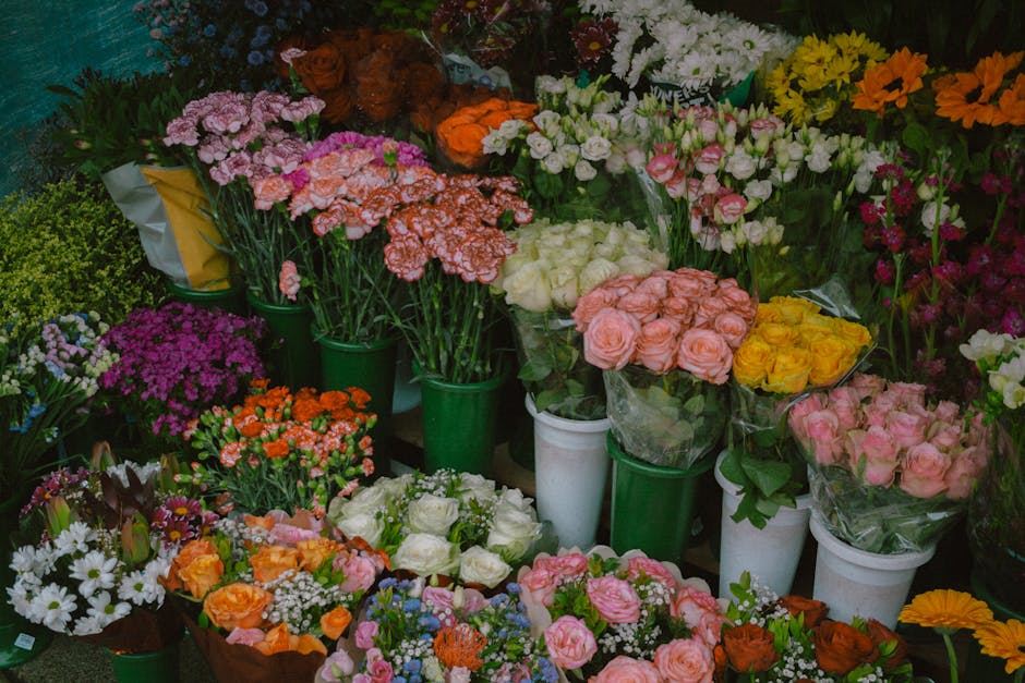 A vibrant display of fresh floral arrangements arranged in green, white, and transparent wrapping, featuring roses in shades of pink, cream, and yellow, as well as carnations, daisies, and other bloom