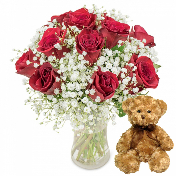 This image portrays a thoughtfully styled romantic gift set, photographed against a perfectly white backdrop so that every petal and stitch is clearly defined. On the left side stands a simple cylindrical glass vase filled with fresh water, holding a generous bouquet of deep crimson red roses. There are about a dozen blooms, some tightly budded, others fully opened to reveal layers of plush, velvety petals that curl softly at the edges. The roses range in tone from a classic true red to slightly darker, wine-like shades near the centre, creating natural depth and dimension in the arrangement. Threaded through the roses are abundant sprays of white baby's breath, each tiny Gypsophila flower like a miniature star suspended on fine green stems. This delicate filler forms a soft, cloud-like aura around the more dramatic roses, making the overall look both luxurious and gentle, much like a bouquet prepared in an Alperton florist's workshop for a Valentine's Day surprise near Stonebridge Park or a quiet anniversary dinner at home. Slender green stems and touches of foliage can be seen through the clear glass, grounding the arrangement in a fresh-from-the-florist realism. To the right of the floral display sits a plush teddy bear in a warm light brown shade, its fur thick and textured, inviting the viewer to imagine its softness. The bear's small round black eyes, embroidered nose, and subtle stitched smile give it an affectionate, almost protective expression, while a dark brown satin ribbon is tied neatly into a bow at its neck. Together, the bouquet and teddy bear convey warmth, love, and celebration, evoking the feeling of a carefully chosen gift delivered to someone special in Alperton.